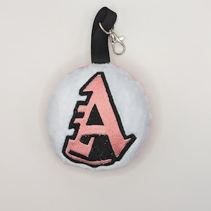 May include: A pink and white plush keychain with a black and pink letter A embroidered on the front. The keychain has a black strap and a silver metal clasp.