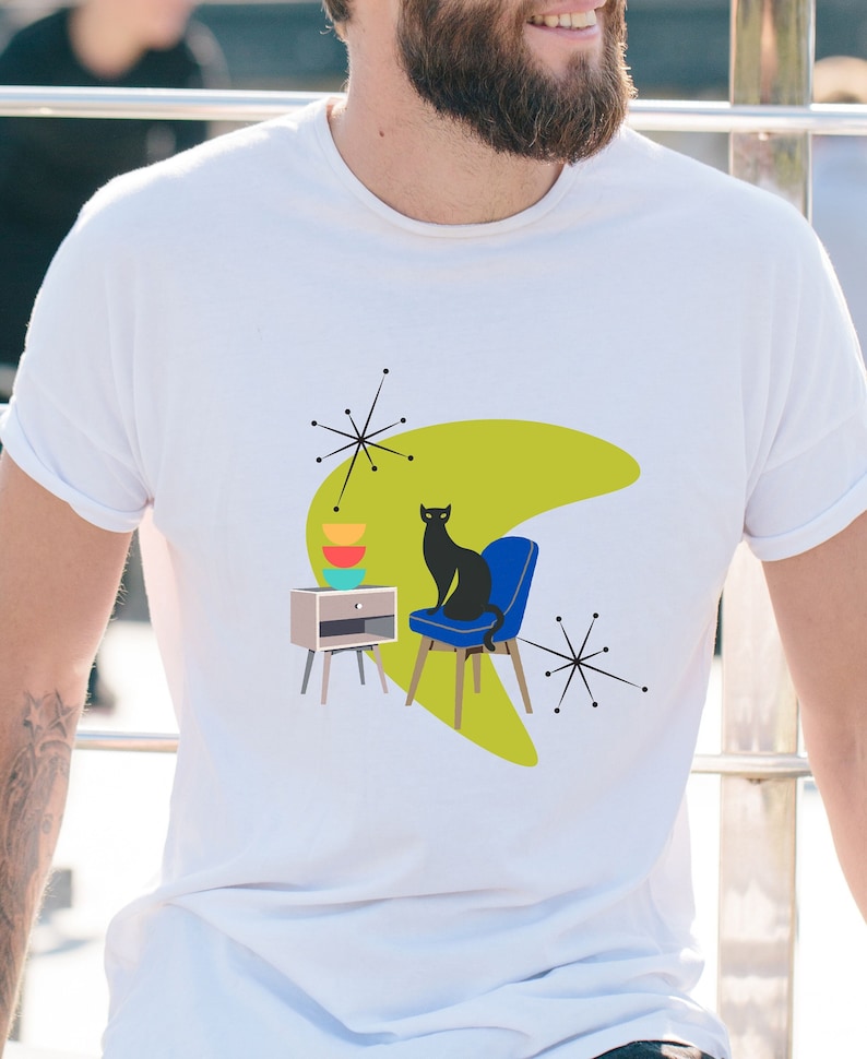 May include: A white t-shirt with a black cat sitting on a blue chair. The cat is in front of a green boomerang shape with a side table and stacked colorful bowls. There are black starbursts on either side of the design.