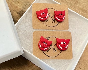 May include: A pair of red pig earrings in a white box with a brown paper insert. The insert has the text "N N" on it.