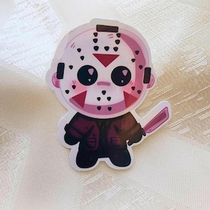 Puede incluir: Una pegatina troquelada que muestra un personaje de dibujos animados con una máscara de hockey blanca con orificios negros para los ojos, marcas triangulares rojas y puntos negros. El personaje viste una chaqueta marrón oscuro y un machete morado.
