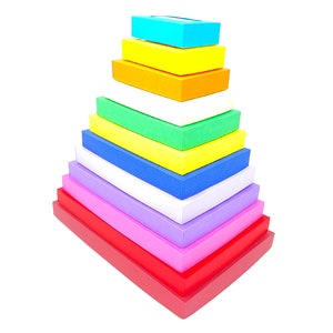 May include: A rainbow-colored stack of rectangular boxes, arranged in a pyramid shape. The boxes are made of a smooth, colorful material and are stacked in order of color, from top to bottom: blue, yellow, orange, white, green, yellow, blue, white, purple, pink, and red.