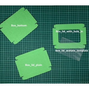 May include: Three green cardboard box templates on a green cutting mat. The templates are labeled "Box_bottom", "Box_lid_plain", and "Box_lid_with_hole_in". A clear acetate template is also visible, labeled "Box_lid_acetate_template".