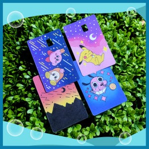 May include: Four rectangular gift tags with cartoon character designs. One tag features Kirby characters, another Pikachu, a third Courage the Cowardly Dog, and the last a sunset scene. Each tag has a small square hole at the top.