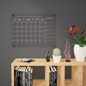 May include: A clear acrylic dry erase calendar with a black background and white lettering. The calendar is mounted on a grey wall. The calendar has a monthly view with the days of the week labelled S, M, T, W, T, F, S. The calendar also has a section for "This Week" with the days of the week labelled S, M, T, W, T, F, S. The calendar is surrounded by a wooden shelf with books and decorative items.