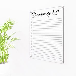 Puede incluir: Tablero de lista de compras de acrílico transparente con las palabras "Shopping List" en escritura negra. El tablero tiene líneas horizontales para escribir y una serie de círculos en el lado izquierdo. El tablero está montado en una pared blanca.