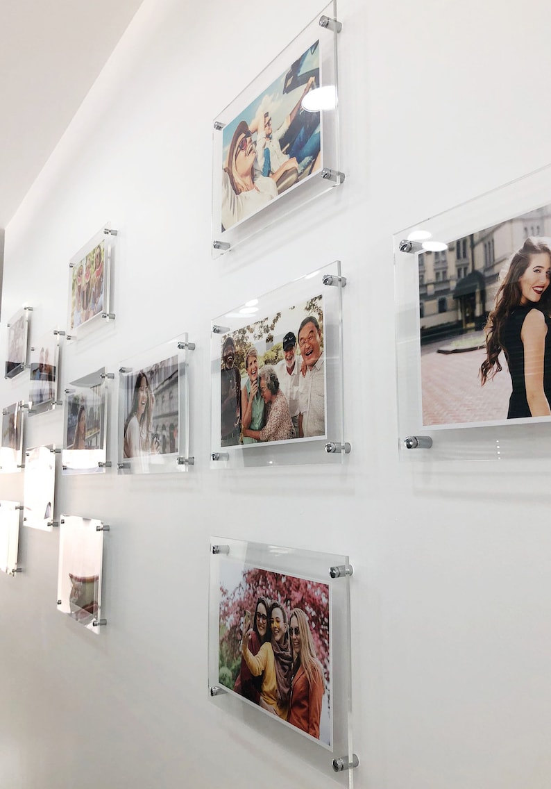 May include: A white wall with multiple framed photos of different people and groups of people. The frames are clear acrylic and have silver metal fasteners.