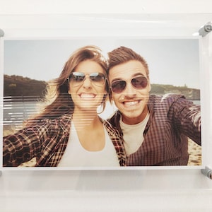 May include: A couple wearing sunglasses smiles for a selfie. The woman is wearing a plaid shirt and the man is wearing a plaid shirt. They are outdoors in a sunny location.