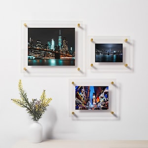 May include: Three acrylic picture frames with gold accents, each displaying a different nighttime cityscape photograph of New York City. The top frame shows the Brooklyn Bridge, the middle frame shows the city skyline, and the bottom frame shows a street scene.