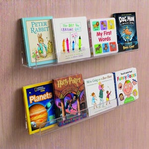 May include: Two clear acrylic shelves mounted on a light wood-grain wall, displaying children's books. Titles include "Peter Rabbit," "The Day the Crayons Quit," "My First Words," "Dog Man," "Planets," "Harry Potter," "We're Going on a Bear Hunt," and "Funny Faces."