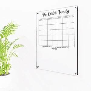 Puede incluir: Un calendario de borrado en seco de acrílico transparente con el texto "The Carter Family" en la parte superior. El calendario tiene espacios para cada día de la semana y cada mes del año. Hay una sección en la parte inferior para notas.