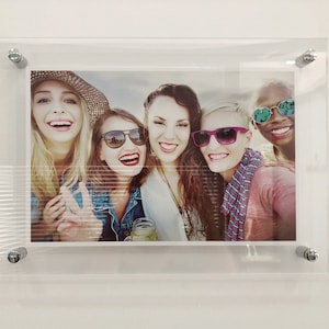 May include: A group of five friends smiling and wearing sunglasses. They are all wearing casual clothing and are standing in front of a blurred background.
