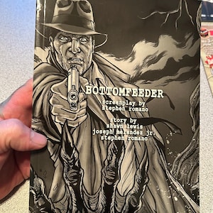 May include: A black and white comic book titled "BOTTOMFEEDER" featuring a man in a fedora pointing a gun. The cover art is a detailed illustration with the title and author credits visible. The comic book is held by a person.