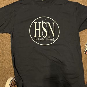 May include: A navy blue t-shirt with a white circular graphic. The graphic features the letters "HSN" inside a circle, with the words "Hail Satan Network" below the letters.