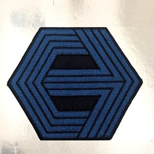 May include: A blue and black embroidered patch in the shape of a hexagon. The patch features a geometric design with multiple layers of lines.