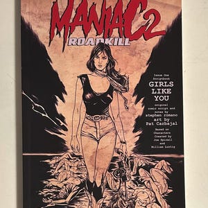May include: Comic book cover titled "MANIAC 2 ROADKILL" featuring a woman in a black top and shorts, holding a camera. The cover art is in a vintage style with the text "GIRLS LIKE YOU".
