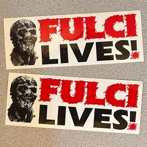 May include: Two white stickers with black and red text. The stickers feature a black and white image of a zombie head and the text "FULCI LIVES!" in red and black.