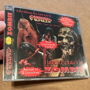 Puede incluir: Una caja de CD con fondo negro y un gráfico rojo y blanco de un zombie. El texto en la caja dice "Umberto Lenzi's Cannibal Ferox", "Make Them Die Slowly!" y "Banned in 31 Countries!"  La caja también presenta una imagen más pequeña de un zombie con el texto "Lucio Fulci's Zombie".