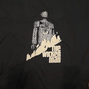 May include: Black t-shirt featuring a white graphic of a wicker man figure engulfed in flames, with the text "THE WICKER MAN" below. The design is a stylized illustration.