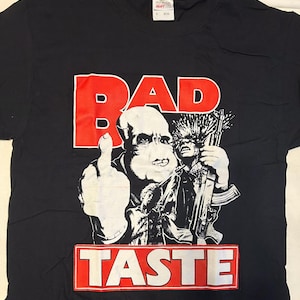 May include: Black t-shirt with a graphic design featuring the word "BAD" in red block letters above a black and white illustration of a person giving the middle finger. The word "TASTE" is below the illustration.