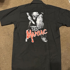 May include: Dark grey short-sleeved shirt with a graphic on the back. The graphic shows a figure holding a knife, with the word "MANIAC" in red. Text above reads "I WARNED YOU NOT TO GO OUT TONIGHT."