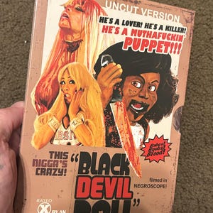 May include: DVD case for the film "Black Devil Doll" with a vintage, horror-movie aesthetic. The cover features illustrations of characters and text including "Uncut Version" and "Babes! Boobs! Blood!" The film is rated X by an all-white jury.