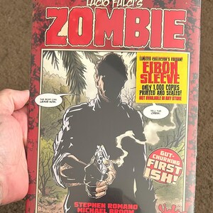 May include: A comic book cover for "Lucio Fulci's Zombie" in red and black. The cover features a man holding a gun, with text that reads "Eibon Sleeve" and "First Ish!" The comic is a limited collector's edition.
