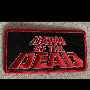 Peut inclure: Un écusson brodé rouge et noir avec le texte "Dawn of the Dead" dans une police de caractères stylisée.