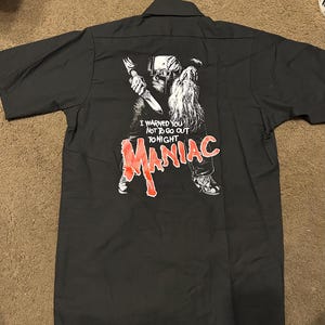 May include: Dark grey short-sleeved shirt with a graphic on the back. The graphic shows a figure holding a knife, with the word "MANIAC" in red. Text above reads "I WARNED YOU NOT TO GO OUT TONIGHT."