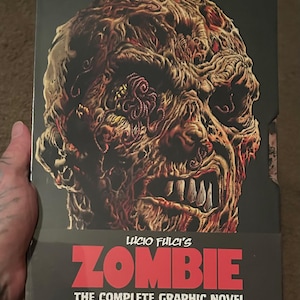 Puede incluir: Caja de novela gráfica con una ilustración detallada de la cara de un zombi. La caja se titula "Lucio Fulci's Zombie: The Complete Graphic Novel" y contiene cuatro libros. La obra de arte muestra un zombi en descomposición con dientes y gusanos expuestos.