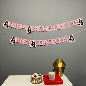 Puede incluir: Una pancarta rosa y blanca con el texto "Happy Bachelorette Baby Gorgeous" e imágenes de mujeres con micrófonos. Un pastel blanco con glaseado de chocolate y una vela sobre una mesa con un jarrón dorado y una alfombra roja.