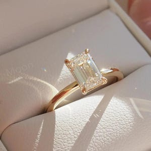 May include: A gold engagement ring with an emerald-cut diamond sits in a white ring box. The ring has a slender, polished band, and the diamond is clear and sparkling. The box is open, and the ring is angled to catch the light.