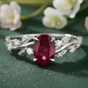 May include: A silver ring with a large oval ruby gemstone. The ring has a leaf and vine design with small, clear accent stones. The background features white flowers and greenery, set against a dark green surface.