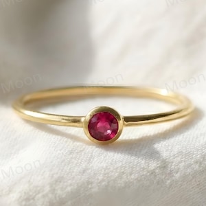 May include: A fine gold ring with a central, circular red gemstone. The ring has a thin band and a bright red stone, creating a simple and elegant design. The ring is set against a soft, white fabric background.