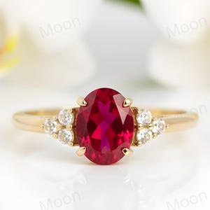 May include: A gold ring featuring a large oval-cut ruby center stone. The ring is flanked by clusters of small, round diamonds on each side. The ring is set against a blurred background of white flowers.