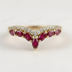 以下が含まれることがあります： V字型のデザインのゴールドトーンのリング。リングには、小さな透明な石の列と、マーキス型の濃い赤色の宝石が並んでいます。リングは白い背景にセットされています。