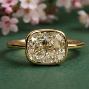 May include: A gold ring with a large, square-cut, light-colored gemstone. The ring is set against a green background with blurred pink and white flowers in the background. The ring's band is thin and simple.