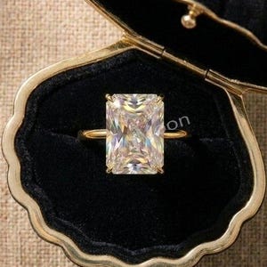 May include: A gold ring featuring a large, rectangular, radiant-cut clear gemstone set in a black velvet, shell-shaped jewellery box with gold-toned trim. The ring is centred within the open box, which displays a soft, dark interior lining.