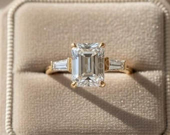 May include: A gold ring with a large, rectangular, clear gemstone as the centerpiece, flanked by two smaller, rectangular gemstones. The ring is set in a velvet ring box.