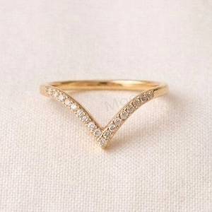 May include: A gold V-shaped ring featuring a series of small, clear, round-cut stones set along the top surface of the band. The metal has a polished finish, and the design forms a pointed chevron shape that tapers towards the centre of the ring.
