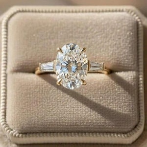 May include: A gold engagement ring with an oval-cut diamond center stone and two baguette diamonds on either side. The ring is set in a gold band and is displayed in a beige velvet ring box.