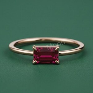 Puede incluir: Un anillo de oro con una gema rectangular de color rojo intenso. El anillo tiene una banda delgada y un diseño sencillo y elegante. La gema es el punto focal, engastada en un estilo minimalista sobre un fondo verde.