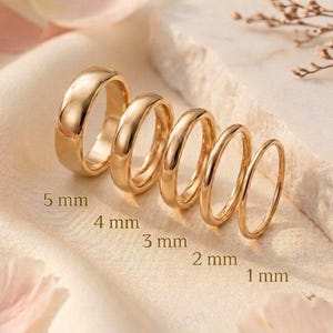 Puede incluir: Cinco anillos de tono dorado dispuestos en fila, disminuyendo en ancho de 5 mm a 1 mm. Cada anillo presenta una superficie lisa y pulida con un perfil redondeado. Los anillos se muestran en secuencia, con el ancho de cada banda claramente indicado.