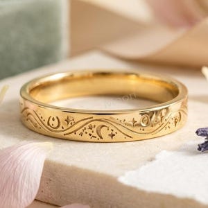 May include: A gold-toned metal ring featuring an engraved celestial pattern that includes crescent moons, stars, and swirling lines. The band has a polished finish and a consistent width, showcasing detailed etchings of various moon phases and small star motifs around the entire circumference of the ring.