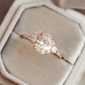 Peut inclure: Une bague en or avec un grand diamant de forme ovale et deux diamants plus petits de chaque côté. La bague est dans un écrin à bague en velours gris.
