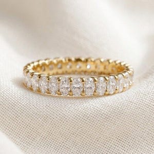 May include: A gold eternity band featuring a continuous row of oval-cut clear stones set in a polished metal frame. The ring displays a repeating pattern of faceted gems around the entire circumference, creating a uniform and sparkling appearance.