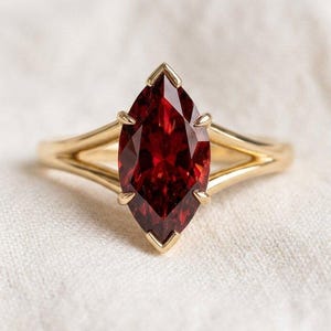 Puede incluir: Un anillo de oro con una gema roja oscura con corte marquesa. La banda del anillo se divide en un diseño de doble banda a cada lado de la gema, creando un aspecto abierto. La gema está engastada con garras puntiagudas.