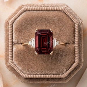 May include: A gold ring featuring a large, rectangular, emerald-cut deep red gemstone as the center stone, flanked by two smaller, rectangular baguette-cut clear stones on each side, all set on a thin, polished gold band.