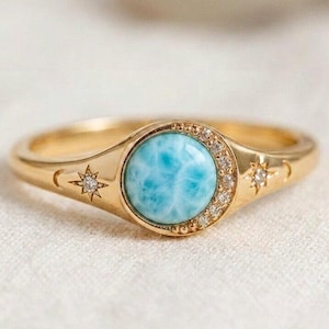 May include: A gold ring featuring a round, light blue gemstone center, surrounded by small clear stones. The ring band has star and crescent moon designs on each side of the gemstone. The ring is set on a white surface.