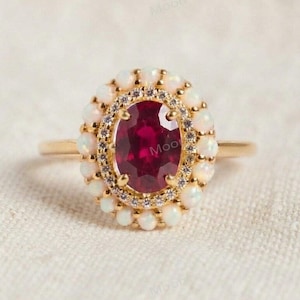May include: A gold ring featuring a central oval-cut deep red gemstone surrounded by a halo of small white opals and a thin border of tiny clear stones. The word Moon is faintly visible in the background behind the center of the ring.