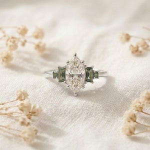 Puede incluir: Un anillo de plata con una gema central transparente de talla marquesa, flanqueada por dos gemas verdes más pequeñas de talla rectangular a cada lado. La banda de metal es pulida y brillante, con un diseño clásico de tres piedras.